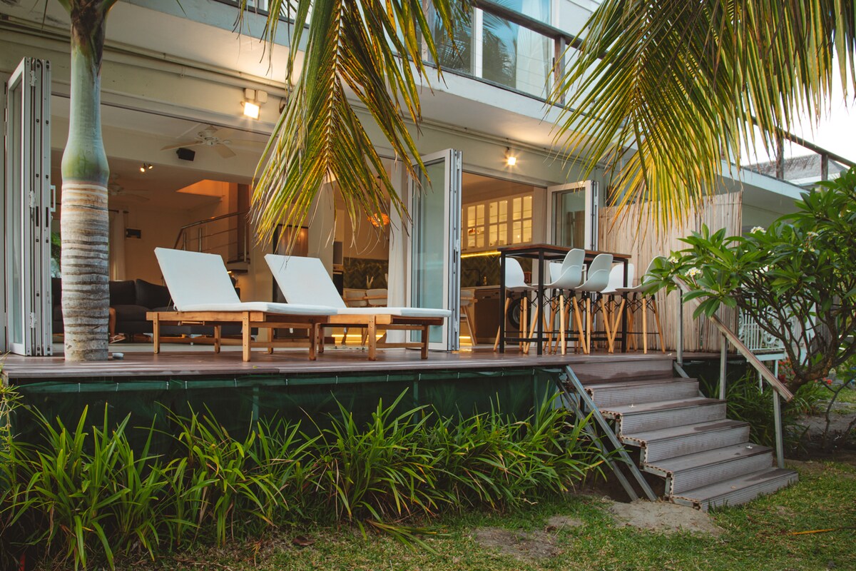 A spacious outdoor area is featured, with wooden sun loungers arranged under swaying palm trees. Steps lead from the lawn to a deck area, where dining furniture is visible. The large glass doors of the interior space invite natural light and provide an open feel.