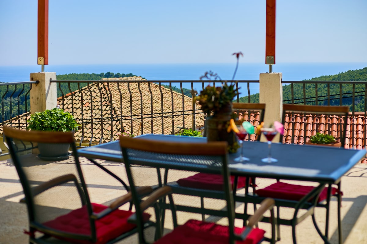 A terrace offers an unobstructed view of the sea and surrounding greenery. A metal table is set with drinks and surrounded by four cushioned chairs. Potted plants add a touch of nature, complementing the tiled roofs below.
