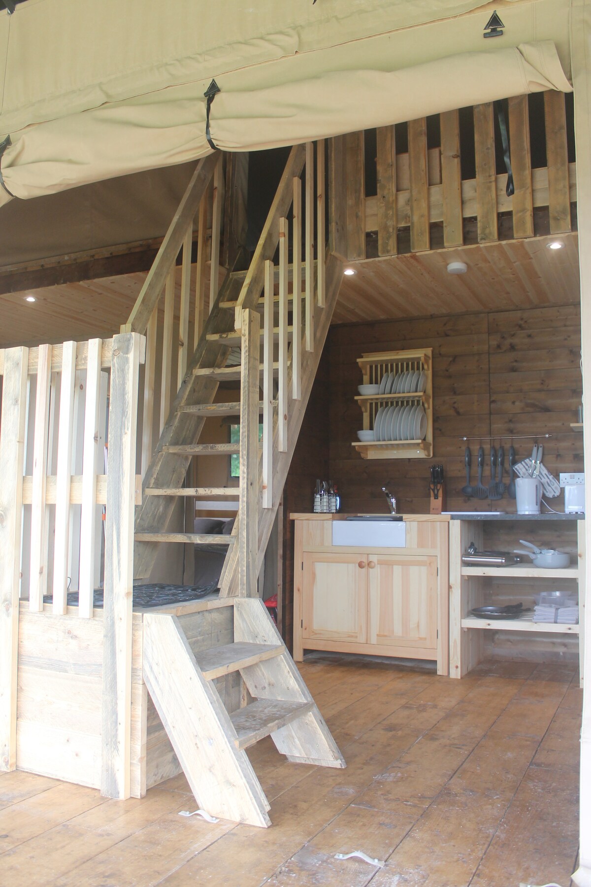 A wooden staircase leads to an upper loft area within the camping tent. Below, a rustic kitchen space features a wooden sink cabinet, neatly arranged dishes, and kitchen utensils displayed on a shelf. The warm wood flooring complements the natural ambiance.
