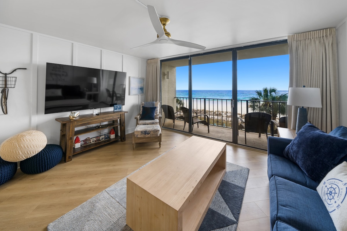 A stylish living area showcases a large flat-screen television mounted on a white wall, accompanied by a wooden coffee table and a cozy blue sofa. Floor-to-ceiling glass doors offer a scenic view of the ocean and access to a private balcony with outdoor seating.