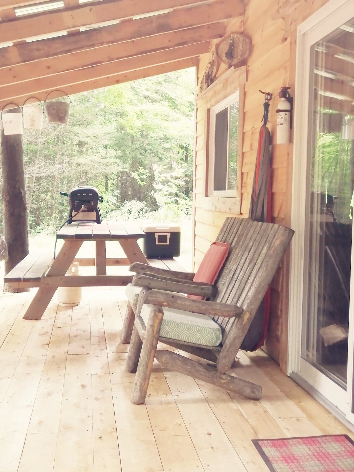 A porch area features a wooden chair with cushions and a picnic table. The table is equipped with a grill. Natural wood tones and a green forest backdrop create a calming outdoor setting, inviting relaxation in a rustic atmosphere.