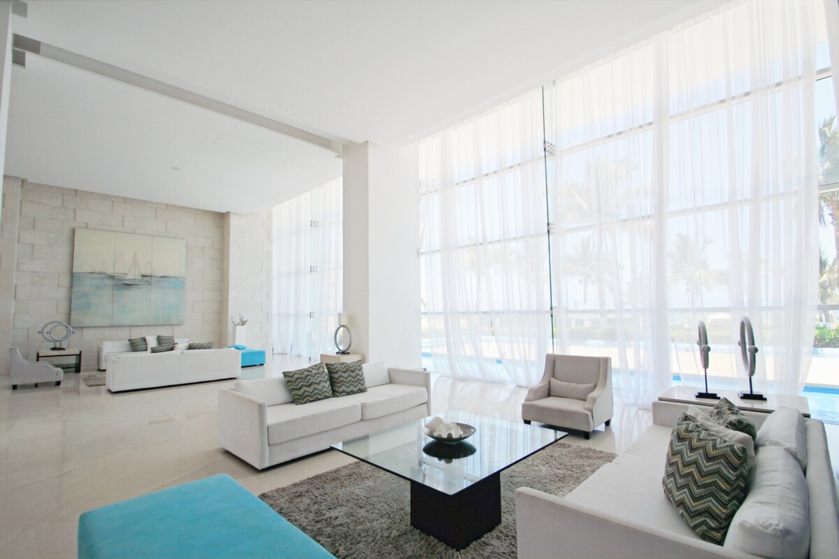 A bright living area features floor-to-ceiling windows allowing natural light to fill the space. Modern white sofas are arranged around a glass coffee table, complemented by decorative cushions. A large abstract painting adds a touch of elegance to the light-colored walls.