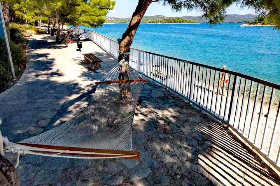 A relaxing area is shown along a waterfront, featuring a hammock positioned between trees. The stone pathway leads to the beach, where sandy shores meet calm blue waters. A railing provides safety along the edge, while benches offer seating for enjoying the view.