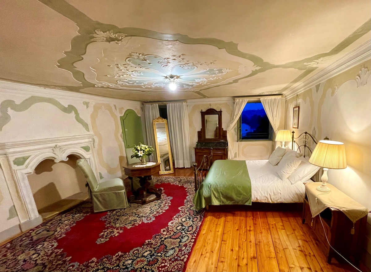 A spacious bedroom showcases elegant frescoed ceilings and soft green accents. A large bed is neatly dressed, flanked by two nightstands with lamps. A vintage desk and chair are positioned by a window, with a cozy area rug and a decorative fireplace enhancing the warmth of the space.