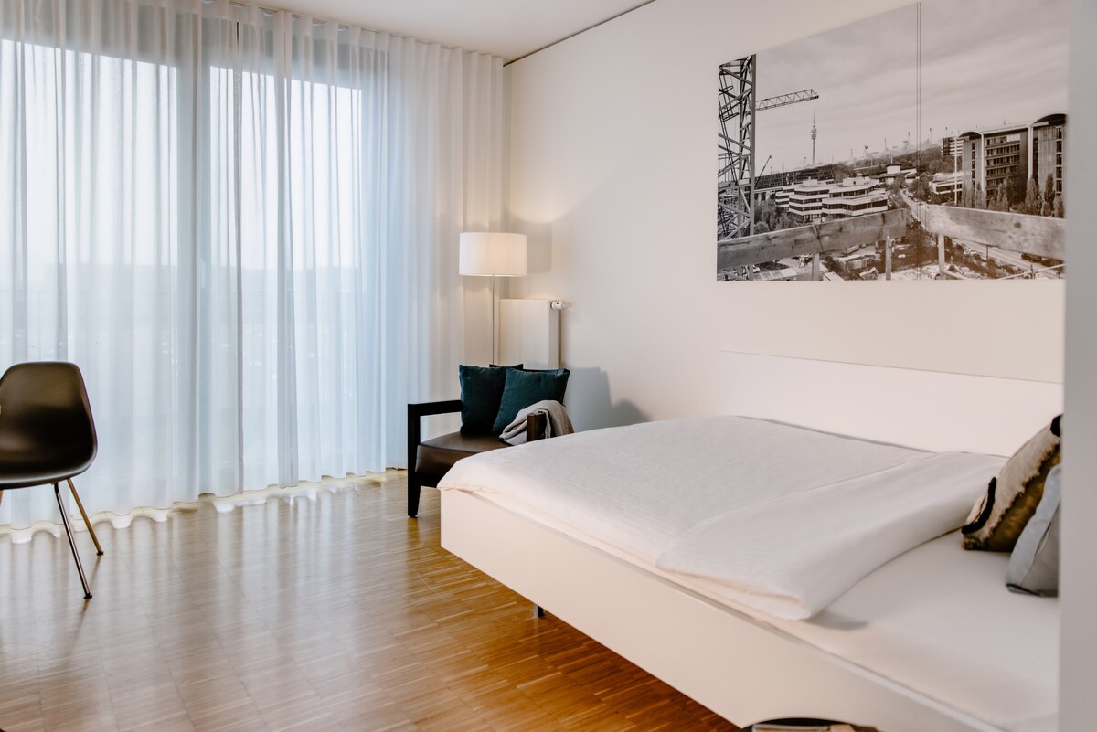 A bright room features a spacious bed with crisp bedding. Natural light filters through sheer curtains, creating a serene atmosphere. A black chair and a side table are positioned nearby, while a large black-and-white photograph decorates the wall, adding a modern touch.