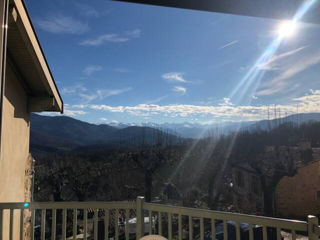 A panoramic view of the mountains is captured from a balcony, showcasing a clear blue sky and distant peaks. Sunlight glimmers as it reflects off the landscape, creating a serene atmosphere. The surrounding area features leafless trees and gentle slopes, emphasizing the natural beauty.