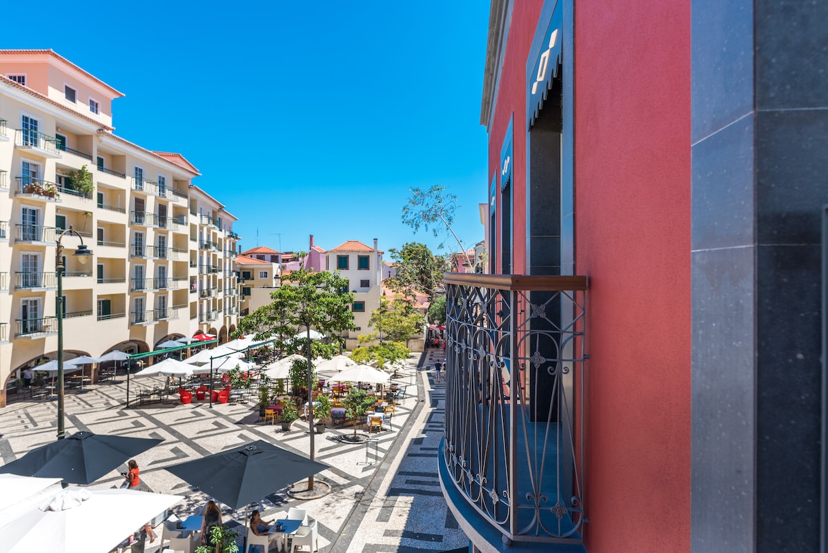 A vibrant plaza is visible, featuring outdoor seating under large umbrellas. Surrounding buildings with balconies create a lively atmosphere, while a clear blue sky enhances the scene. A mix of green trees and colorful chairs adds to the visual appeal of the area.