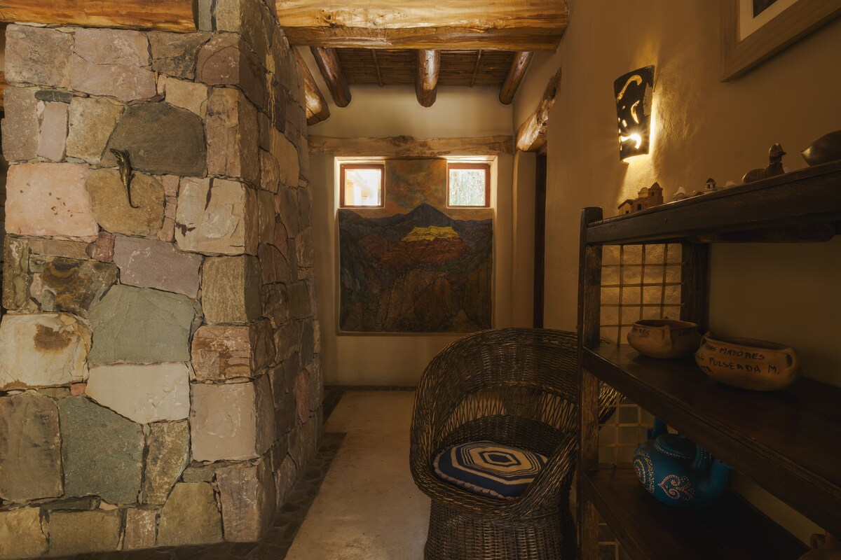 A cozy entryway features natural stone walls and wooden beams, providing a rustic ambiance. A comfortable wicker chair is positioned near a decorative wooden shelf displaying handcrafted items. Two windows allow natural light to illuminate a colorful wall tapestry depicting a scenic landscape.