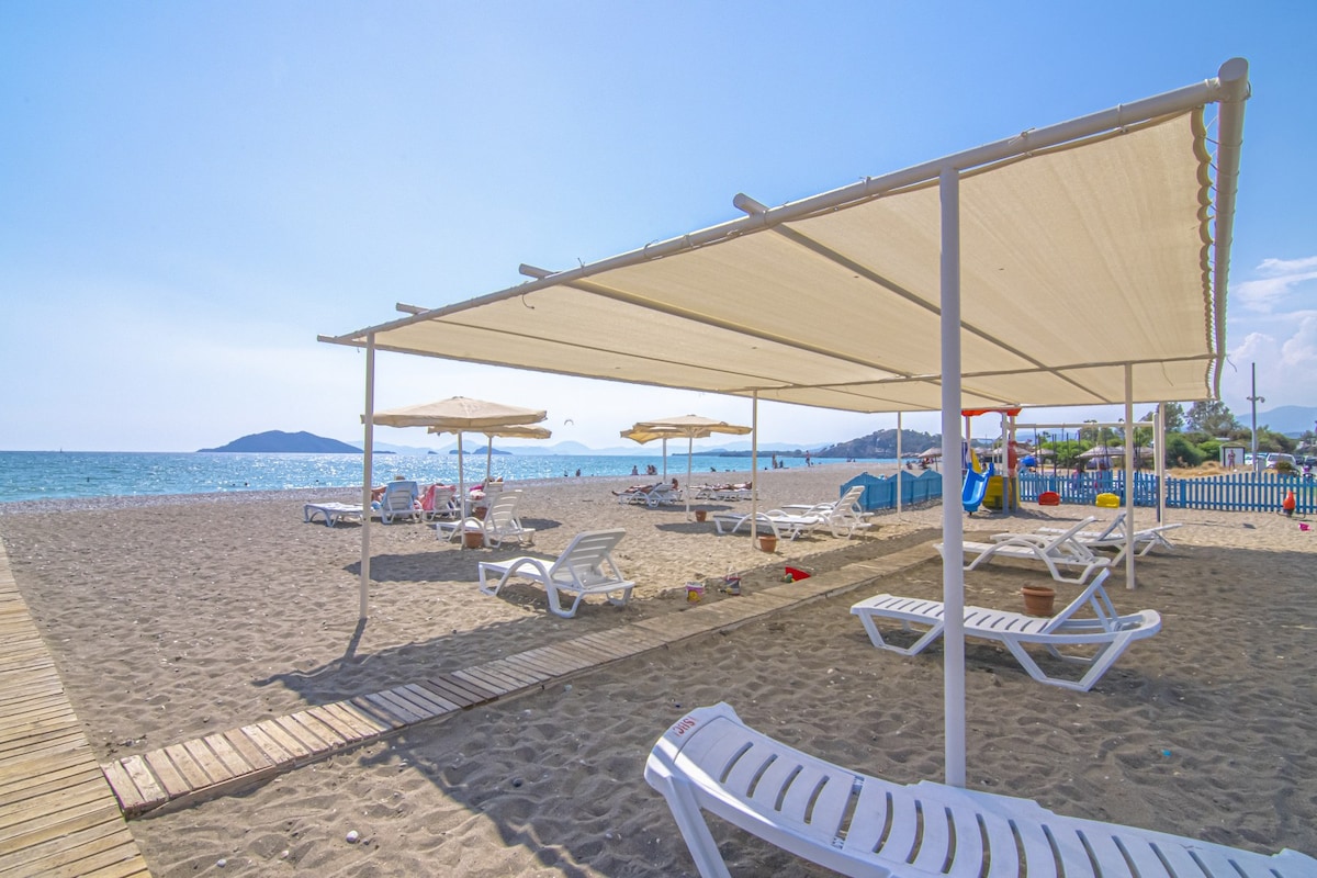 A sandy beach area features rows of lounge chairs under fabric canopies for shade. The calm sea is visible in the background, with umbrellas and recreational areas observed nearby. A wooden pathway leads towards the beach, enhancing accessibility.