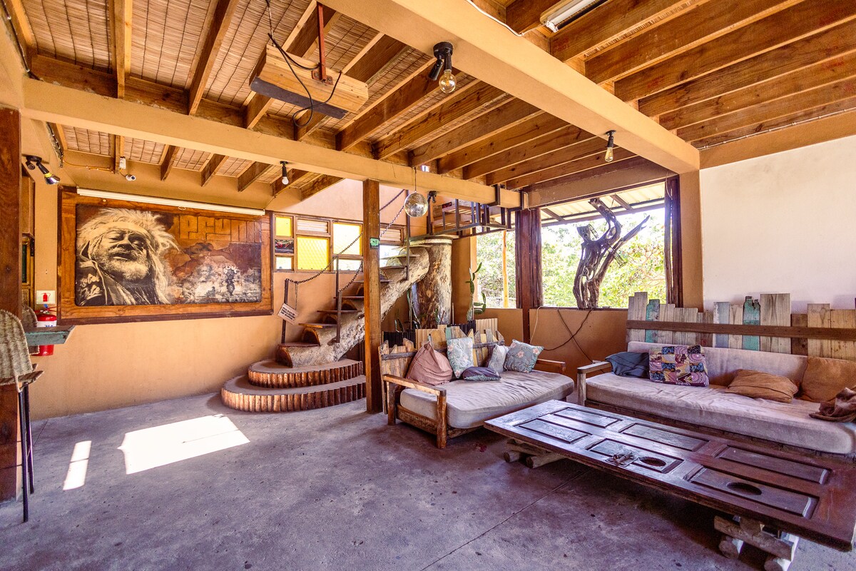 A spacious communal area features a mix of comfortable seating, including large cushions on light-colored sofas. Natural light streams through large windows, and a unique wooden staircase leads to an upper level. The walls display local artwork, contributing to a relaxed atmosphere.