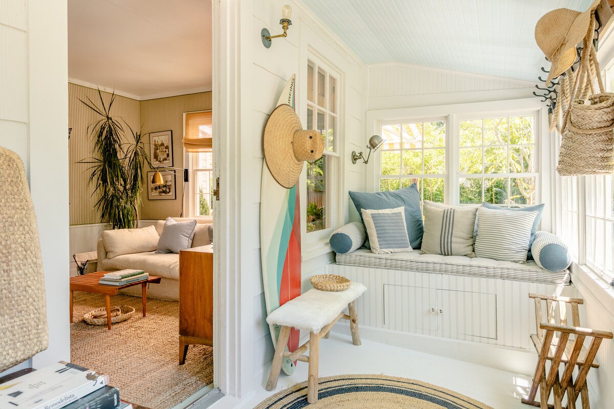 A charming sunroom is showcased with large windows bringing in natural light. A cozy window seat is adorned with blue and white pillows. Nearby, a small wooden table and stool are present, while a glimpse of the inviting living room is visible through the open doorway.