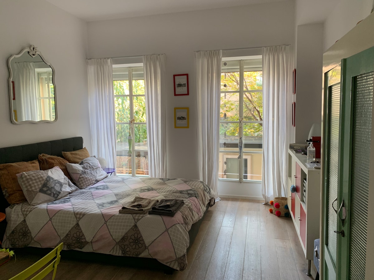 A bright studio is featured with a spacious bed adorned in a patterned quilt. Natural light flows through two windows, draped with sheer white curtains. A small kitchenette is visible on the right, with a mirror reflecting the warm ambiance.