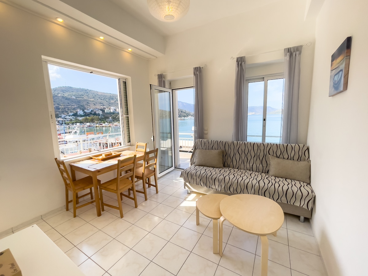 A well-lit living space features a comfortable sofa and a small coffee table. A dining area with wooden chairs is positioned near large windows that offer a view of the scenic waterfront and mountains. Light curtains soften the sunlight filtering into the room.