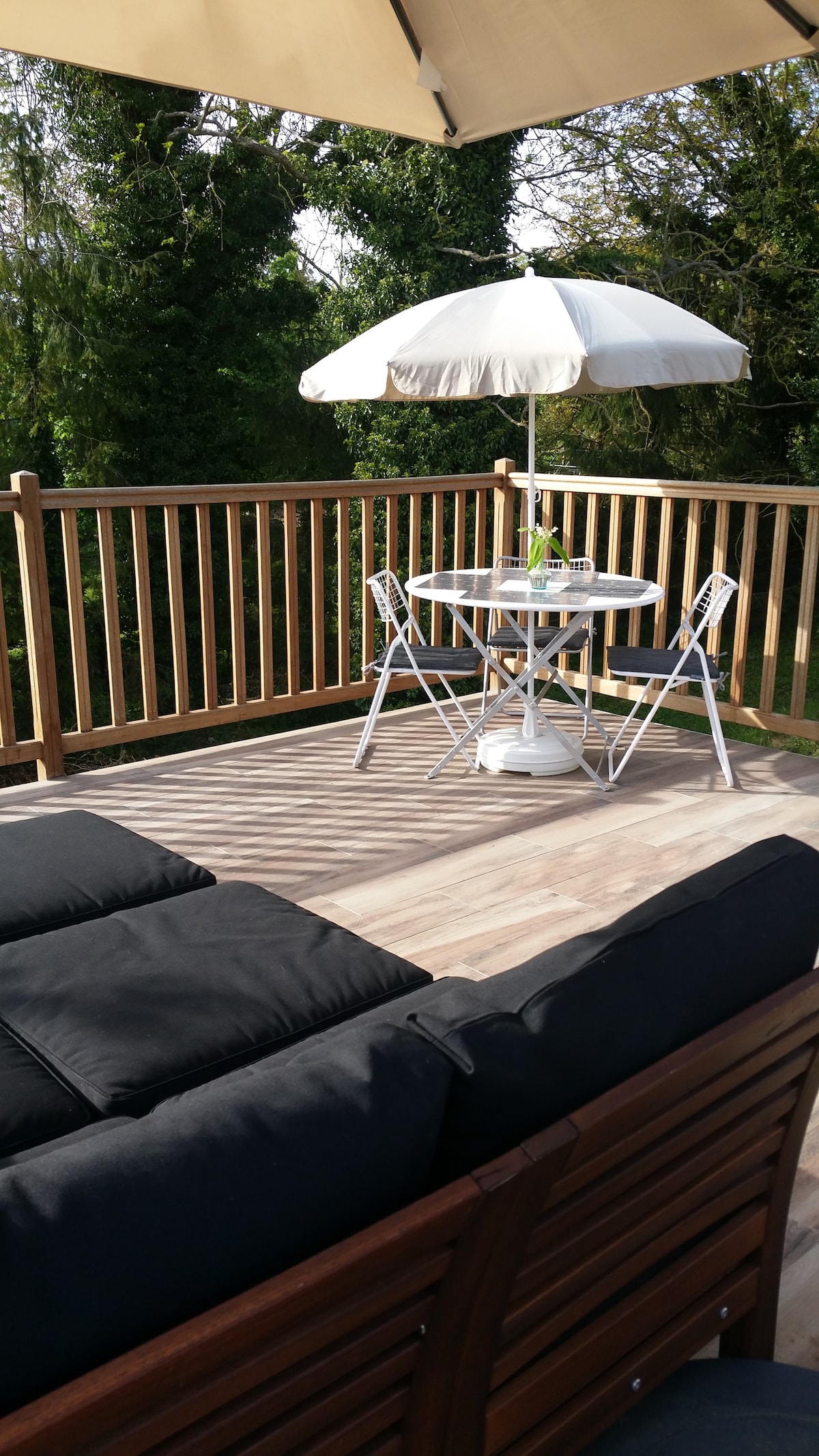 A private terrace features a round dining table with a large umbrella, surrounded by four white chairs. A comfortable black sofa is positioned nearby, providing a cozy seating area. The wooden railing offers a peaceful view of the surrounding greenery.