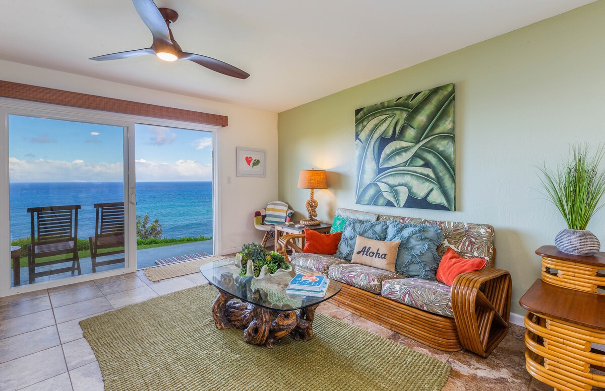 The living area features a comfortable couch adorned with colorful cushions, alongside a glass coffee table that displays a welcome booklet. Large sliding doors reveal stunning ocean views and provide access to outdoor seating, while a vibrant tropical-themed wall art enhances the ambiance.