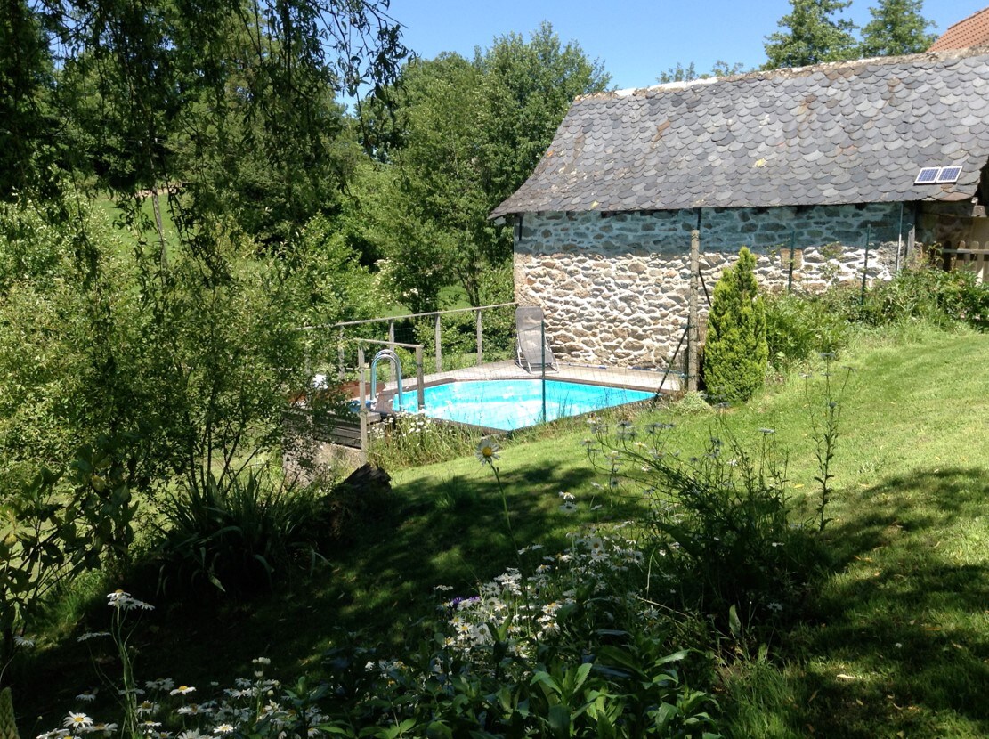 A private swimming pool is set within a lush garden, surrounded by greenery and mature trees. The pool area features a wooden deck, and a stone building is visible in the background. The serene landscape invites relaxation in a natural setting.