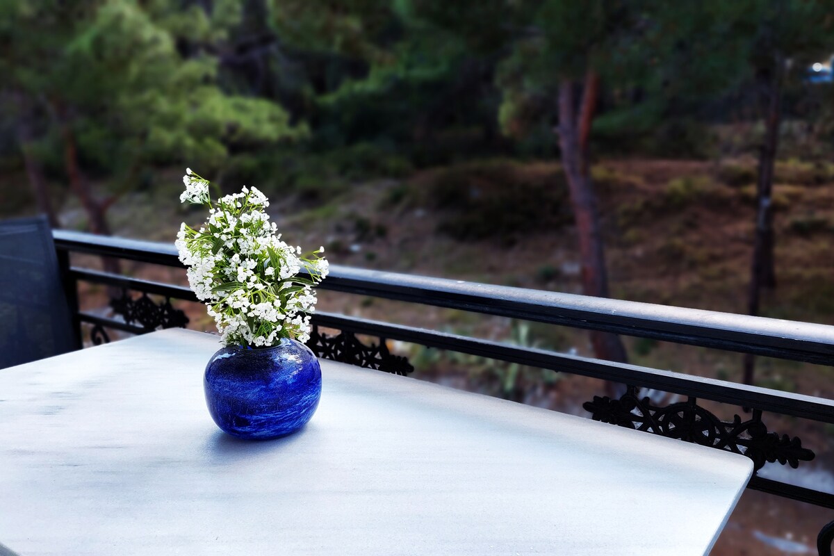 A table is depicted on a balcony, adorned with a blue vase containing fresh white flowers. The background features a serene view of green trees and natural foliage, offering a peaceful ambiance.