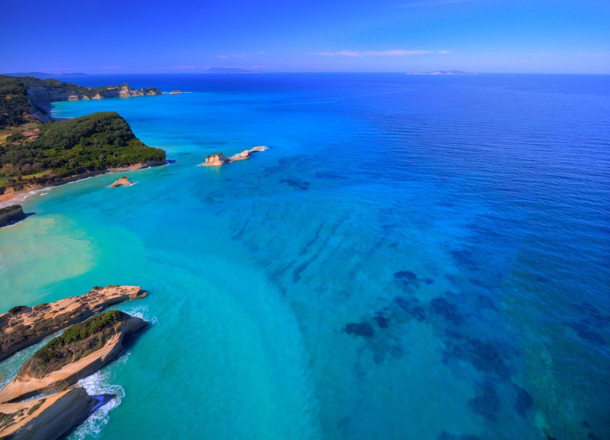 A panoramic view of the coastline reveals tranquil turquoise waters blending into deeper blue tones. Gentle waves lap at the sandy shores, while rocky formations emerge from the sea. The horizon features distant landmasses under a clear blue sky, creating a serene coastal landscape.