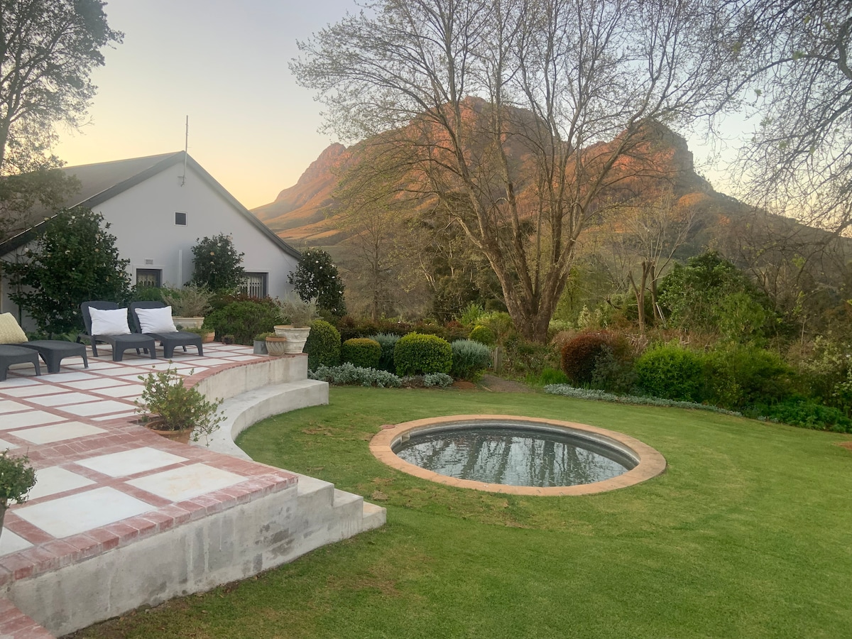 A serene outdoor space features a circular plunge pool surrounded by a well-maintained lawn and lush greenery. Loungers are arranged on a brick terrace, providing a relaxing area to enjoy views of the mountains and trees in the background.