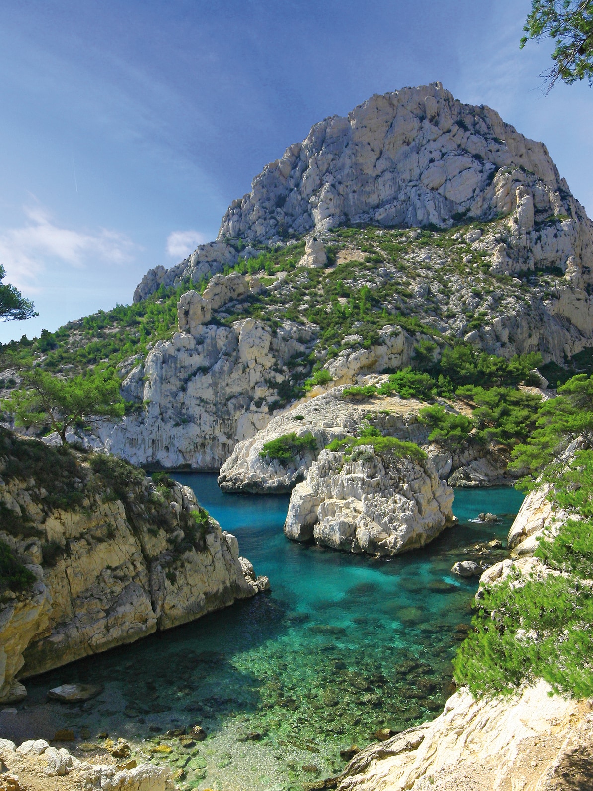 A serene coastal landscape is presented, highlighting a clear blue water cove surrounded by rocky cliffs and lush greenery. Towering mountains rise in the background, reflecting the vibrant colors of the sea and sky, creating a harmonious natural setting.