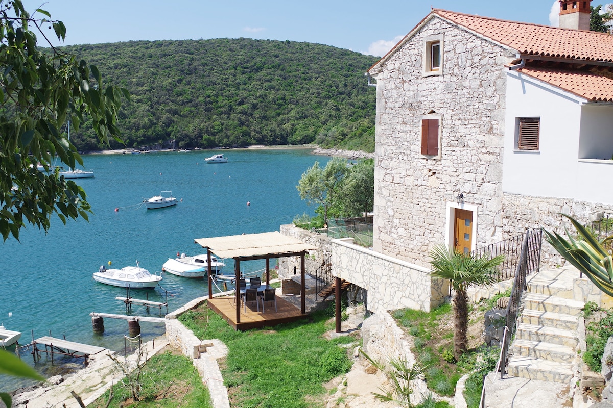 A traditional stone house is positioned near a calm bay, with boats gently bobbing on the water. A wooden gazebo is visible at the water's edge, surrounded by greenery, providing a shaded area to relax. The landscape includes smooth pathways leading down to the water.