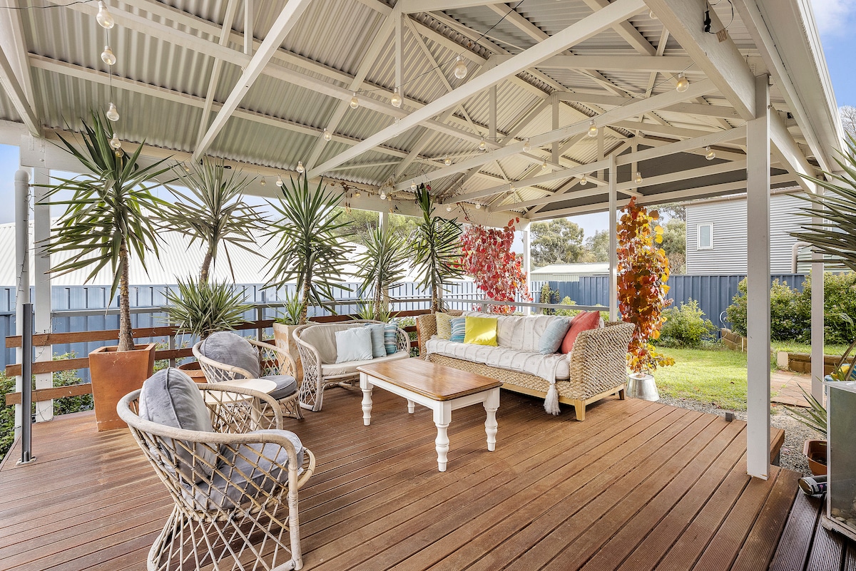 A covered outdoor deck area contains a white wicker sofa, three armchairs, and a wooden coffee table. Potted plants and colorful foliage enhance the space. The deck overlooks a garden area with manicured greenery and a wooden fence, inviting relaxation and outdoor enjoyment.