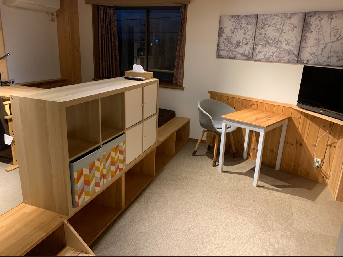 A functional living space is depicted, featuring a wooden shelving unit with a colorful design. A small desk and a chair are positioned nearby, alongside a wall-mounted television. Natural light enters through a window, complemented by soft curtains and decorative wall art.