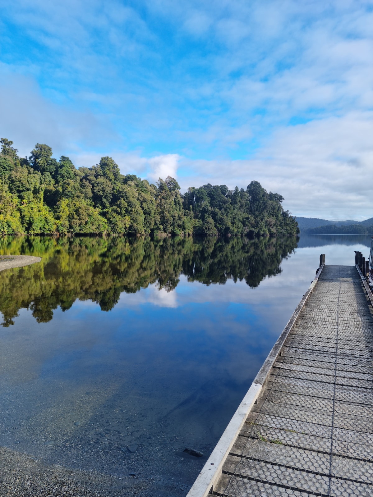 A wooden pier extends over a calm body of water, reflecting the lush greenery of surrounding trees. The sky features a blend of soft clouds and blue hues, creating a serene atmosphere that invites relaxation.
