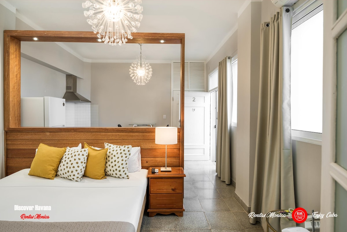 A comfortable sleeping area features a spacious bed adorned with yellow accent pillows and a textured throw. A wooden bedside table is positioned next to the bed. The space is well-lit with natural light streaming through windows beside the bed.