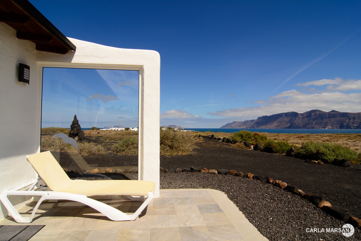 A shaded lounge chair is positioned on a stone patio, offering a direct view of the ocean and nearby cliffs. A large glass window reveals the expansive landscape, framed by the modern white exterior of the building. Natural stone elements complement the setting.