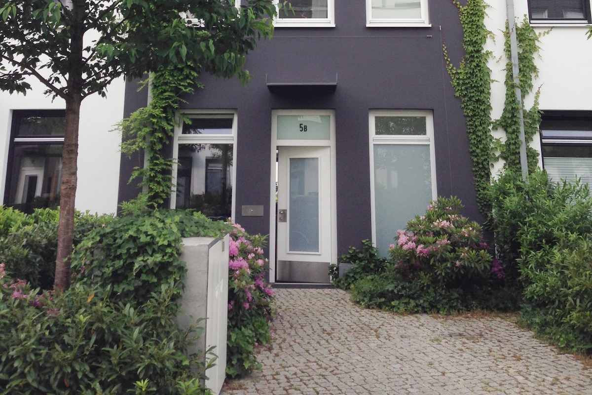 The front entrance of a modern townhouse features a sleek glass door framed by lush greenery and flowering plants. Smooth cobblestones form the path leading to the doorway, complemented by large windows that allow light to penetrate the home's interior.