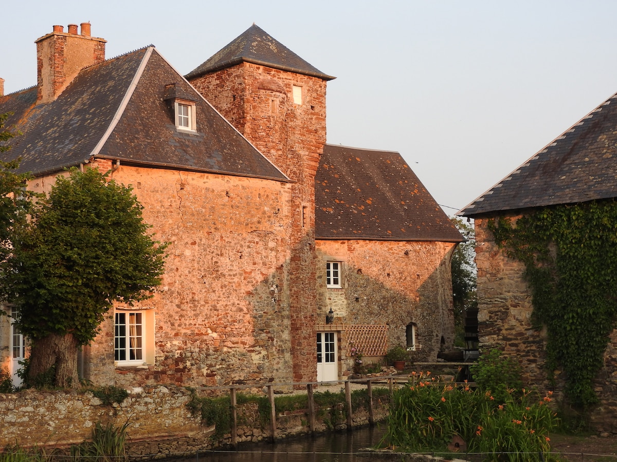 The historic manor is depicted with warm light reflecting off its weathered stone walls. A mix of architectural elements, including a prominent tower and slate roofs, are visible. Lush greenery surrounds the property, contributing to the tranquil ambiance at the water's edge.