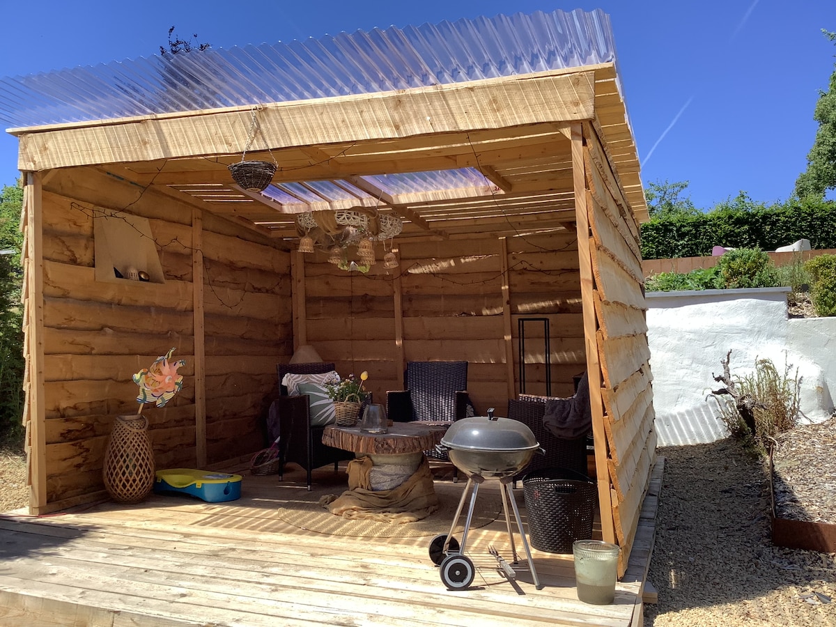 An outdoor wooden structure features a wooden deck with a circular table surrounded by two chairs. A charcoal grill is positioned nearby. The space is illuminated by natural light from the skylight and is adorned with hanging plants, creating a serene and functional area for relaxation.