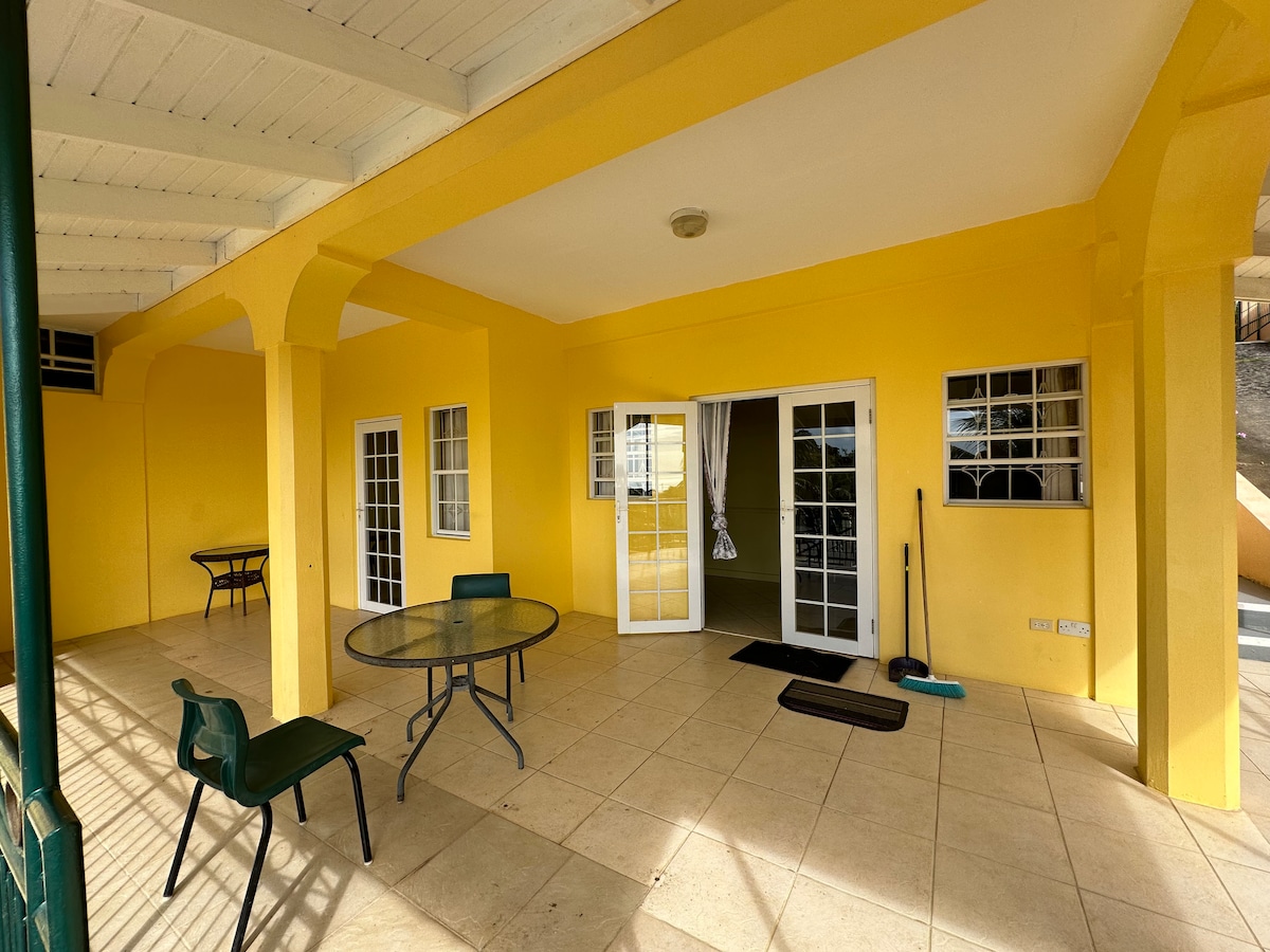 A spacious outdoor patio is featured with a circular glass table and four green chairs. Bright yellow walls create a cheerful backdrop. Large glass doors lead into the interior space, while light-colored tiled flooring is visible. A broom is positioned near the entrance.