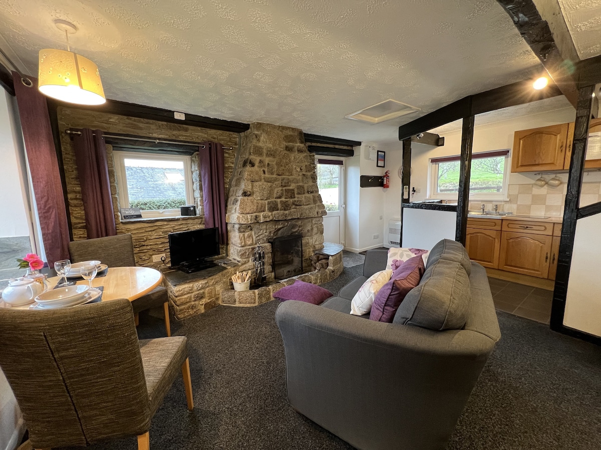 A cozy living area features a stone fireplace as the focal point, surrounded by comfortable seating. A dining table for two is positioned nearby, and an open kitchen is visible in the background. Large windows provide natural light and views of the surrounding countryside.