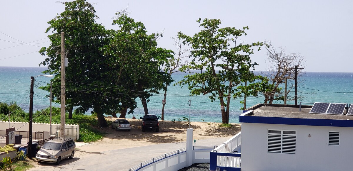 A view of the serene beach and ocean is captured from the balcony. Lush green trees frame the scene, while nearby vehicles are parked along the pathway. The blue waters of the ocean gently meet the shoreline, offering a calming coastal experience.
