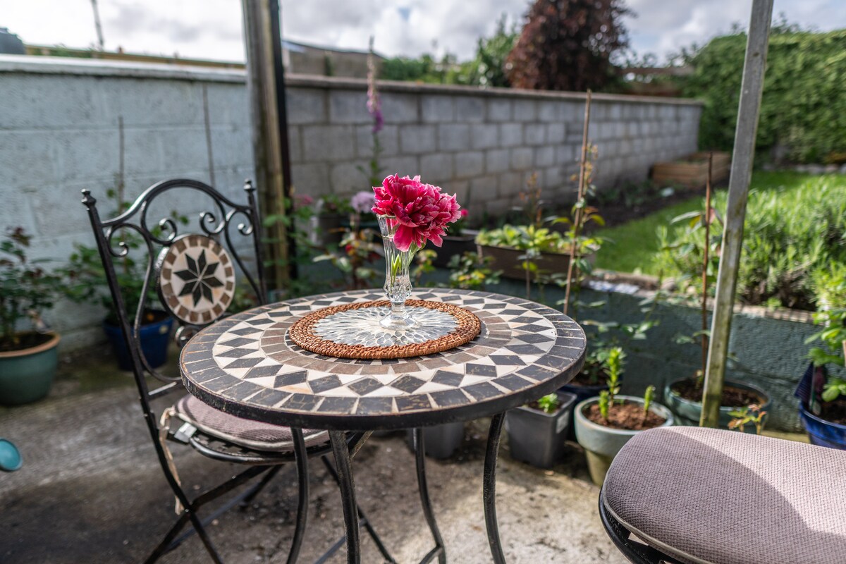 An outdoor setting is depicted with a round table featuring a decorative mosaic top. A small vase with bright flowers is centered on a woven placemat. Surrounding plants are seen in pots, and a chair with a patterned backrest is positioned nearby.