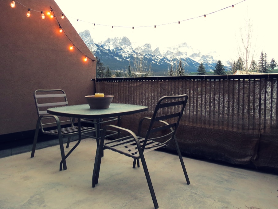A private patio features a table and two chairs, surrounded by a fence. Strung lights offer a soft illumination, with mountains in the background providing a scenic view. The outdoor space is designed for relaxation and enjoying the fresh air.