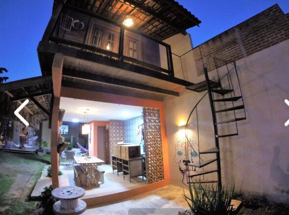 The exterior of the wooden house is shown at dusk, featuring a spiral staircase leading to upper-level balconies. A spacious patio area is visible, with outdoor seating and decorative elements. Soft lighting enhances the welcoming ambiance, surrounded by greenery and inviting pathways.