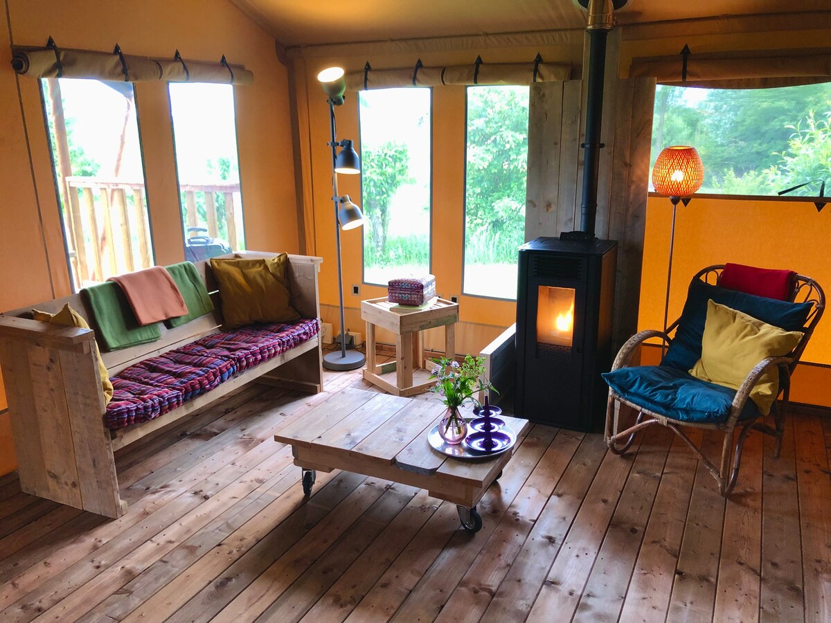 The interior of the safari tent presents a cozy seating area with a wooden sofa and a colorful woven blanket. A small wooden table is positioned beside a charming stove, while large windows allow natural light to fill the space, surrounded by greenery.