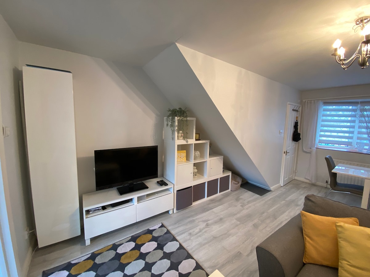A cozy lounge area is featured with a corner sofa and a television. Storage units with a mix of open and closed compartments are positioned alongside a window that brings in natural light. A dining table is visible in the background, against a gently sloped wall.