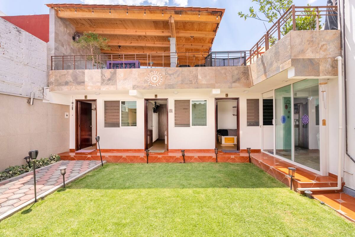 An outdoor terrace is featured, bordered by a well-maintained lawn. The structure showcases a combination of stone and wood, with multiple entrances leading to separate rooms. Ample natural light brightens the space, complemented by areas for seating and relaxation.