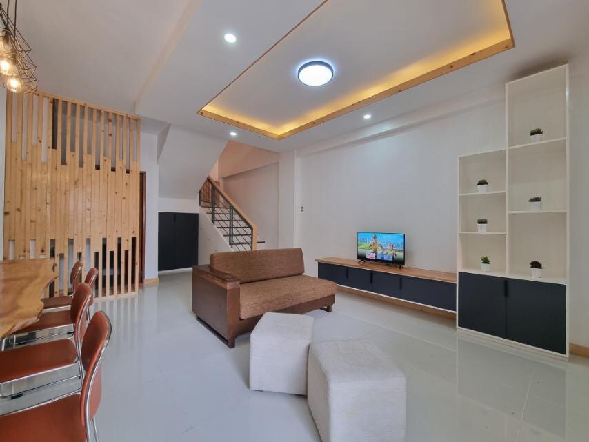 A spacious living area features a brown sofa facing a media unit with a television displaying a scenic view. Modern decor includes wooden accents and light fixtures. The open layout connects to a dining area, with a table and chairs visible, enhancing the communal atmosphere.