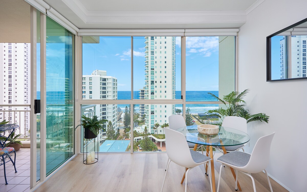 A dining area features a glass table surrounded by white chairs, positioned next to floor-to-ceiling windows. The ocean views extend to the horizon, with glimpses of the coastline and landscaped resort grounds visible below.