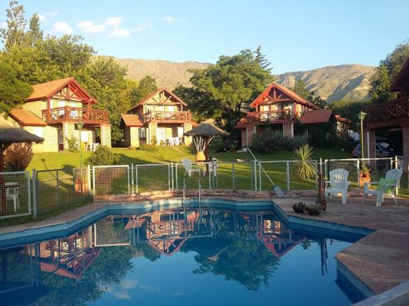 A serene outdoor area features a sparkling pool surrounded by lounge chairs and a grassy landscape. Four charming cabins, each with a balcony, are visible in the background under a clear blue sky, framed by trees and mountains.