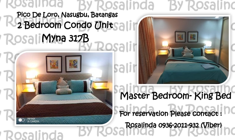 The master bedroom features a large king bed with a soft blue quilt and coordinated pillows. Two bedside lamps provide ambient lighting beside the bed. Neutral wall colors and artwork add visual interest, while warm light filters through the curtains.