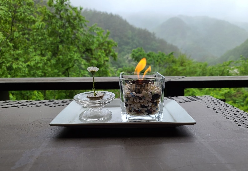 A glass candle holder filled with pebbles features a small flame, resting on a white plate. Beside it, a delicate flower is presented in a transparent dish. Lush green mountains can be seen in the background, partially shrouded in mist.