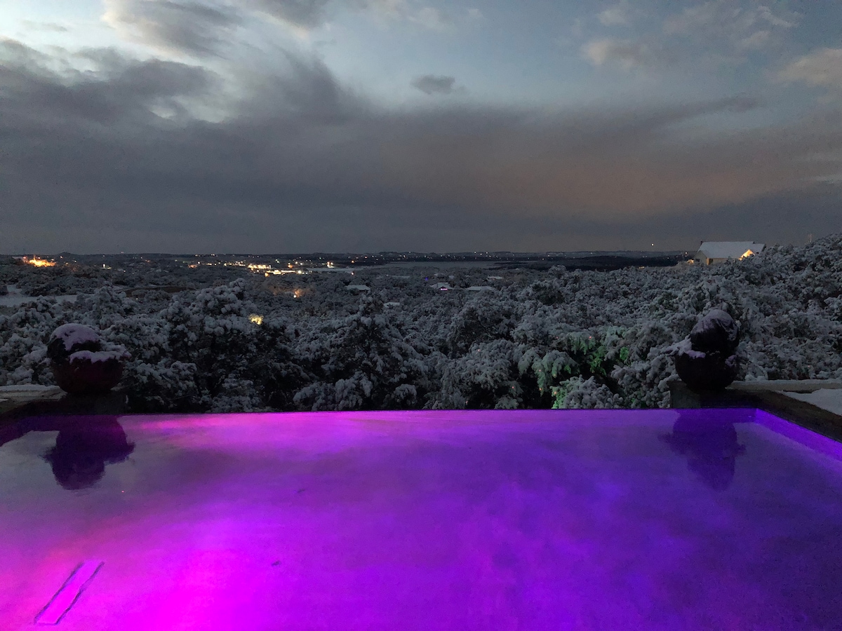 The infinity edge pool appears tranquil, illuminated by soft purple lighting. A sweeping view of the snow-dusted landscape stretches into the distance, with twinkling lights from nearby areas contrasting the serene colors of dusk in the sky.