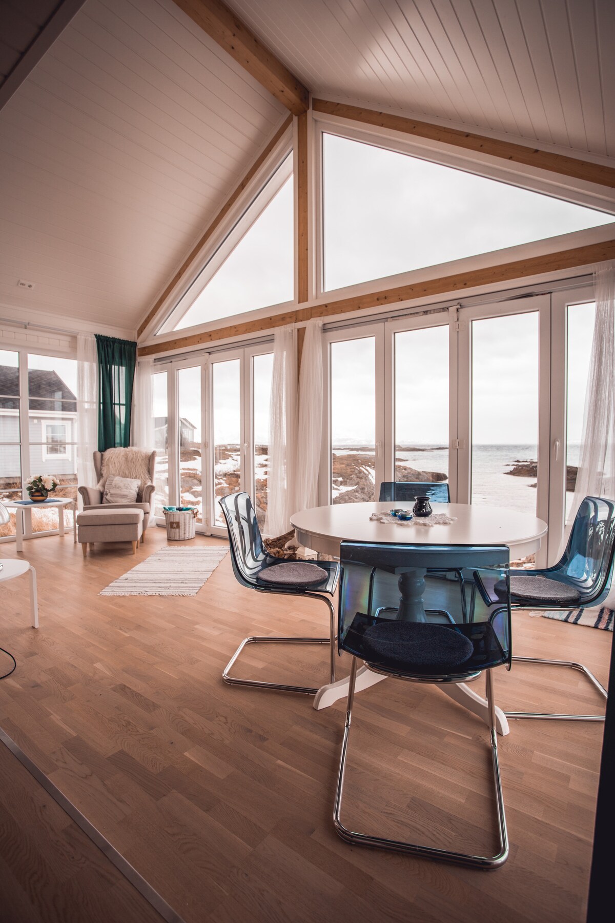 An open living and dining area features large glass doors that allow natural light to fill the space, showcasing stunning views of the outdoors. A round dining table is complemented by transparent chairs, and a cozy seating area with a plush armchair is visible in the corner.