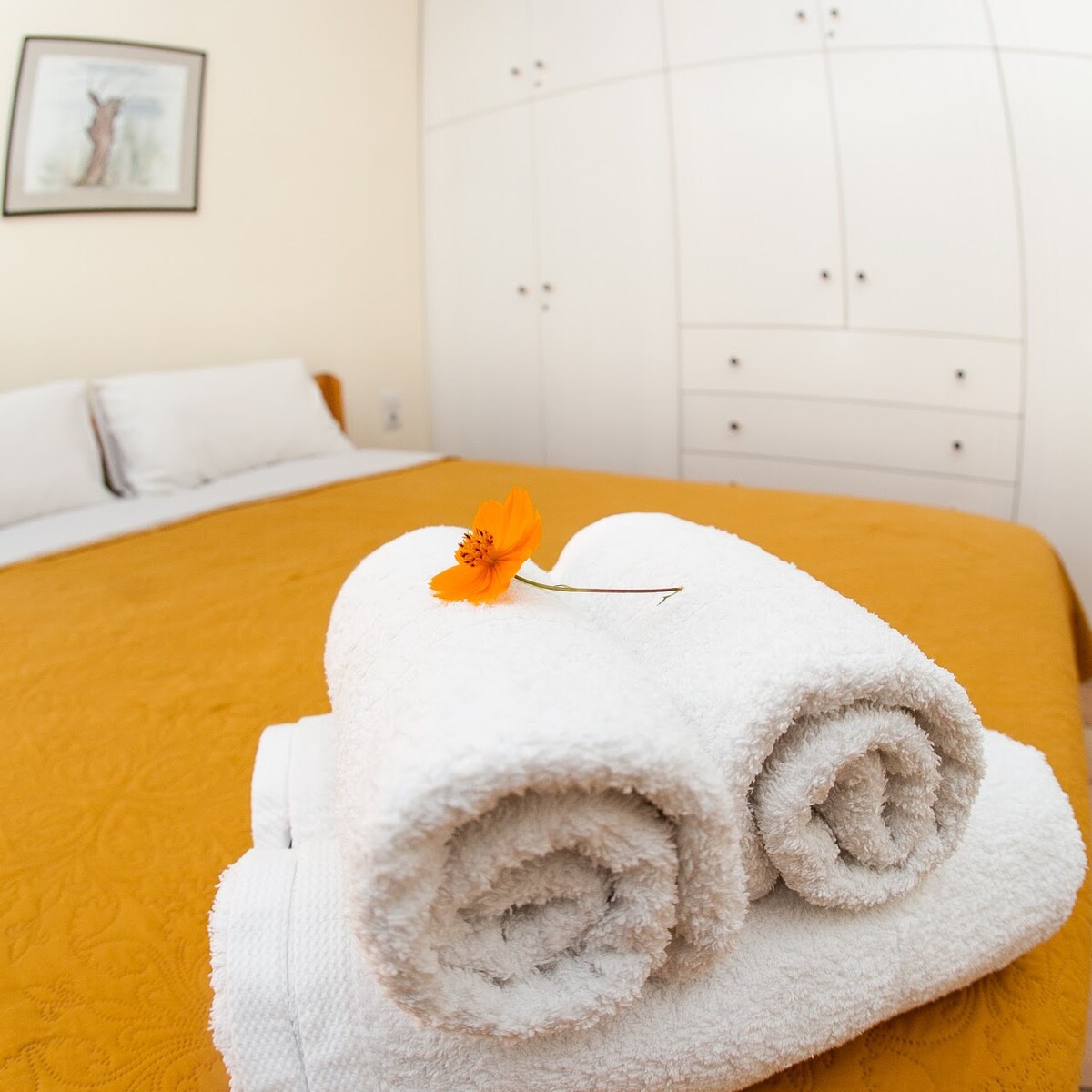 White towels are neatly rolled and placed on a bed with an ochre-colored bedspread. A single orange flower rests atop the towels. The bedroom features soft lighting and a framed artwork is visible on the wall, enhancing the peaceful ambiance.
